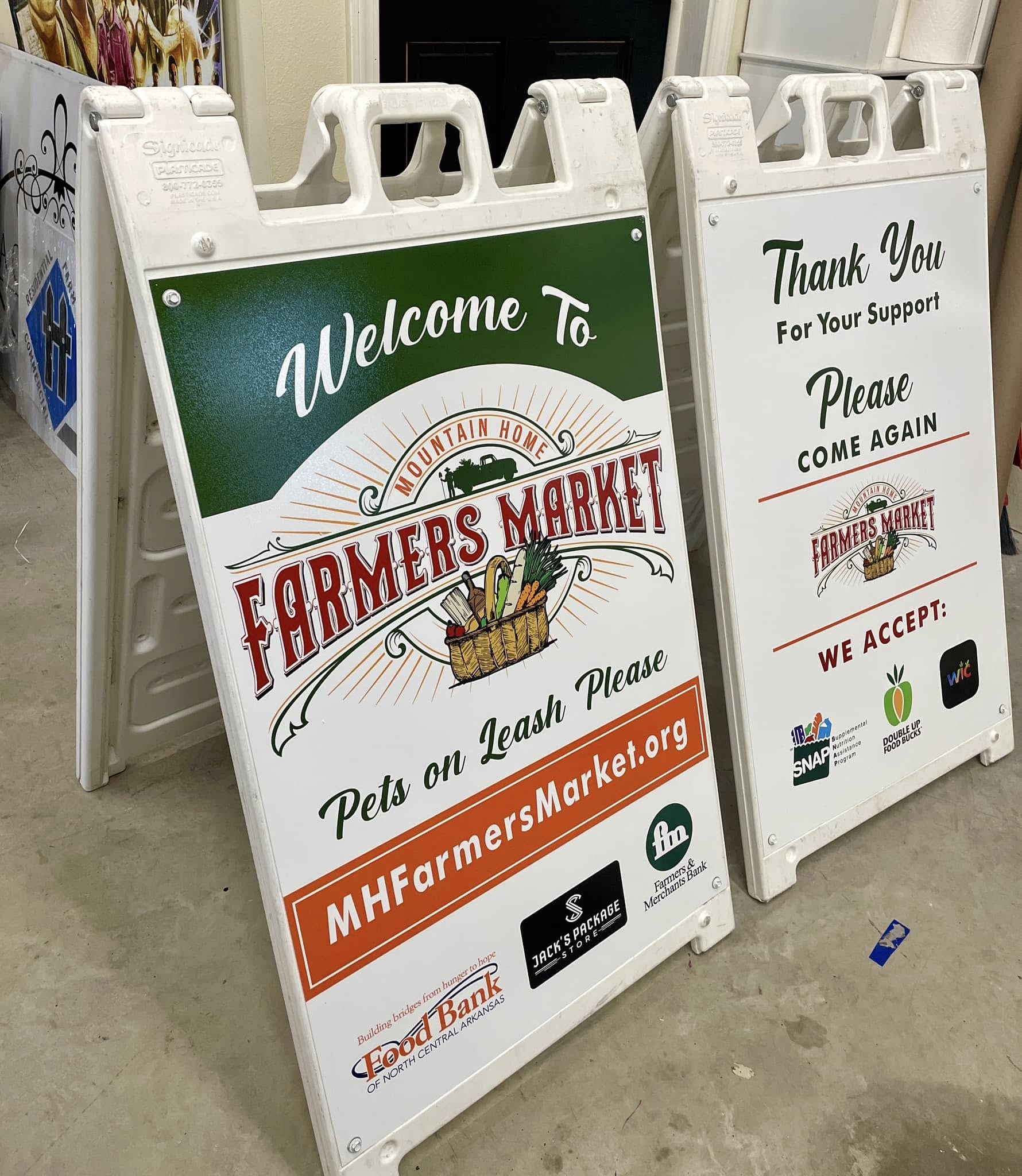 Two white A-frame signs with green and orange Mountain Home Farmers Market graphics.