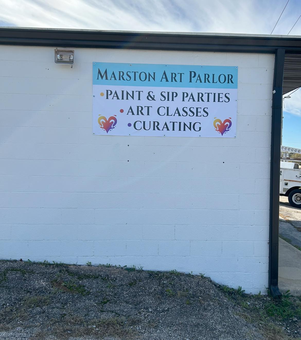 Vinyl banner for Marston Art Parlor on a white brick wall, listing services like 'Paint & Sip Parties'