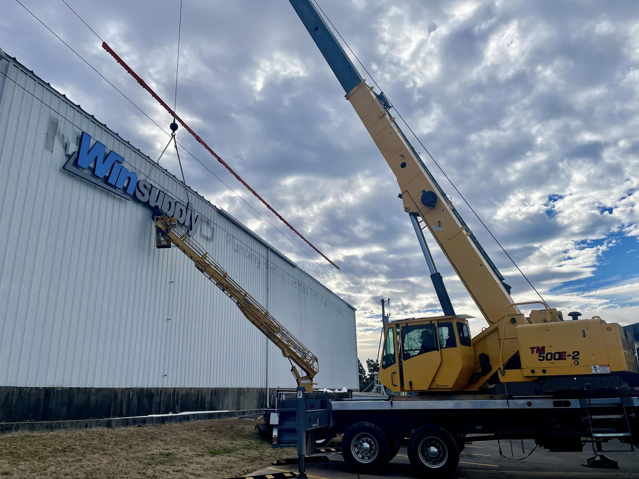 Blue channel letters spelling 'Winsupply' being installed on a white building fascia by a construction crane.