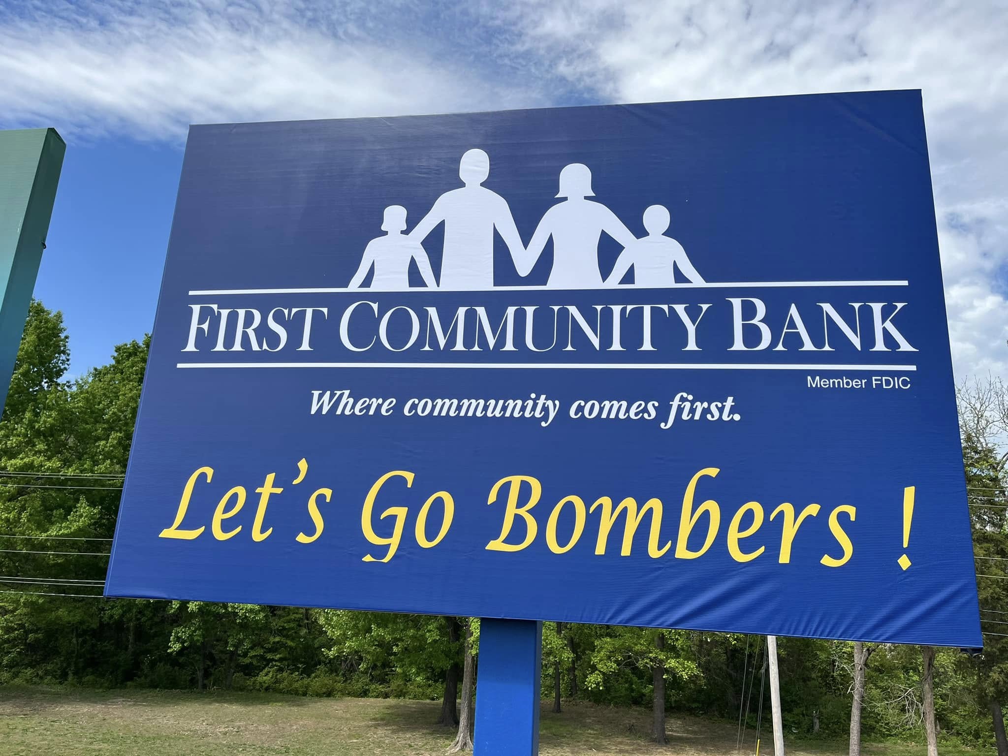 Large blue pylon sign for First Community Bank with a family logo and 'Let's Go Bombers !' in yellow.