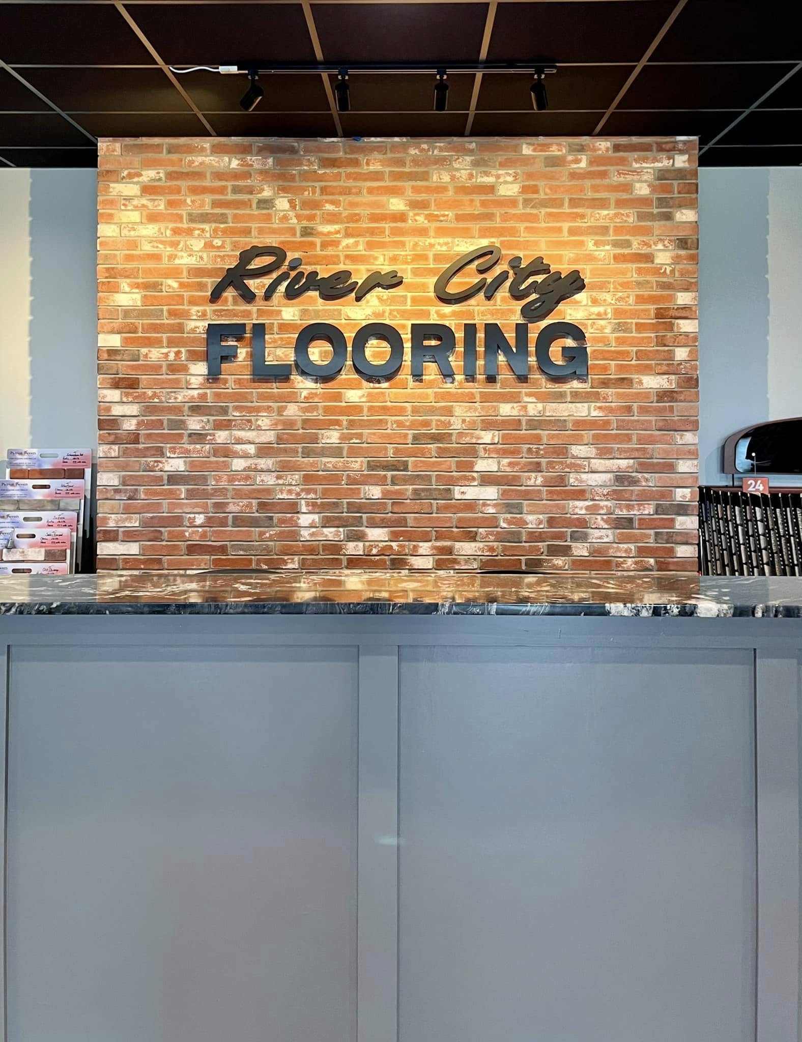 Black dimensional letters spelling "River City Flooring" on an interior brick wall with track lighting.