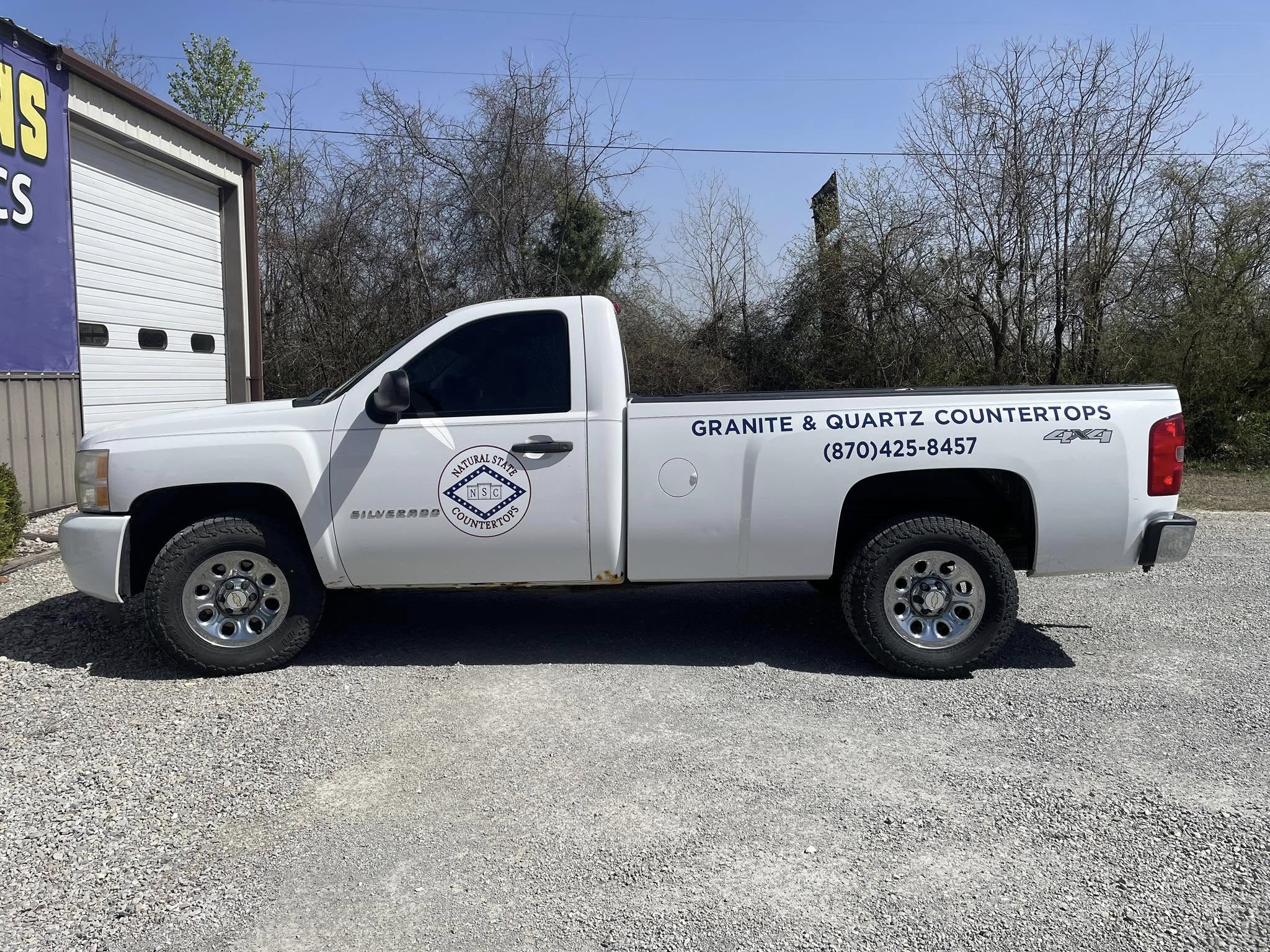 White GMC Sierra pickup truck with blue vinyl text and diamond logo for 'Natural State Countertops' on sides.