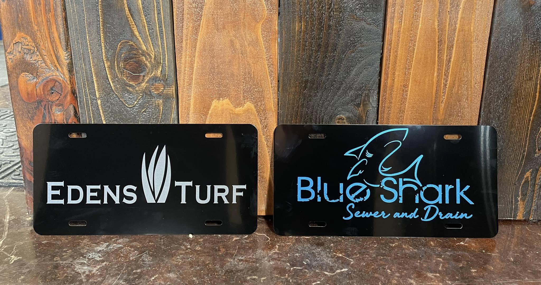 Custom black license plates with white and blue vinyl logos for Edens Turf and Blue Shark Sewer & Drain.
