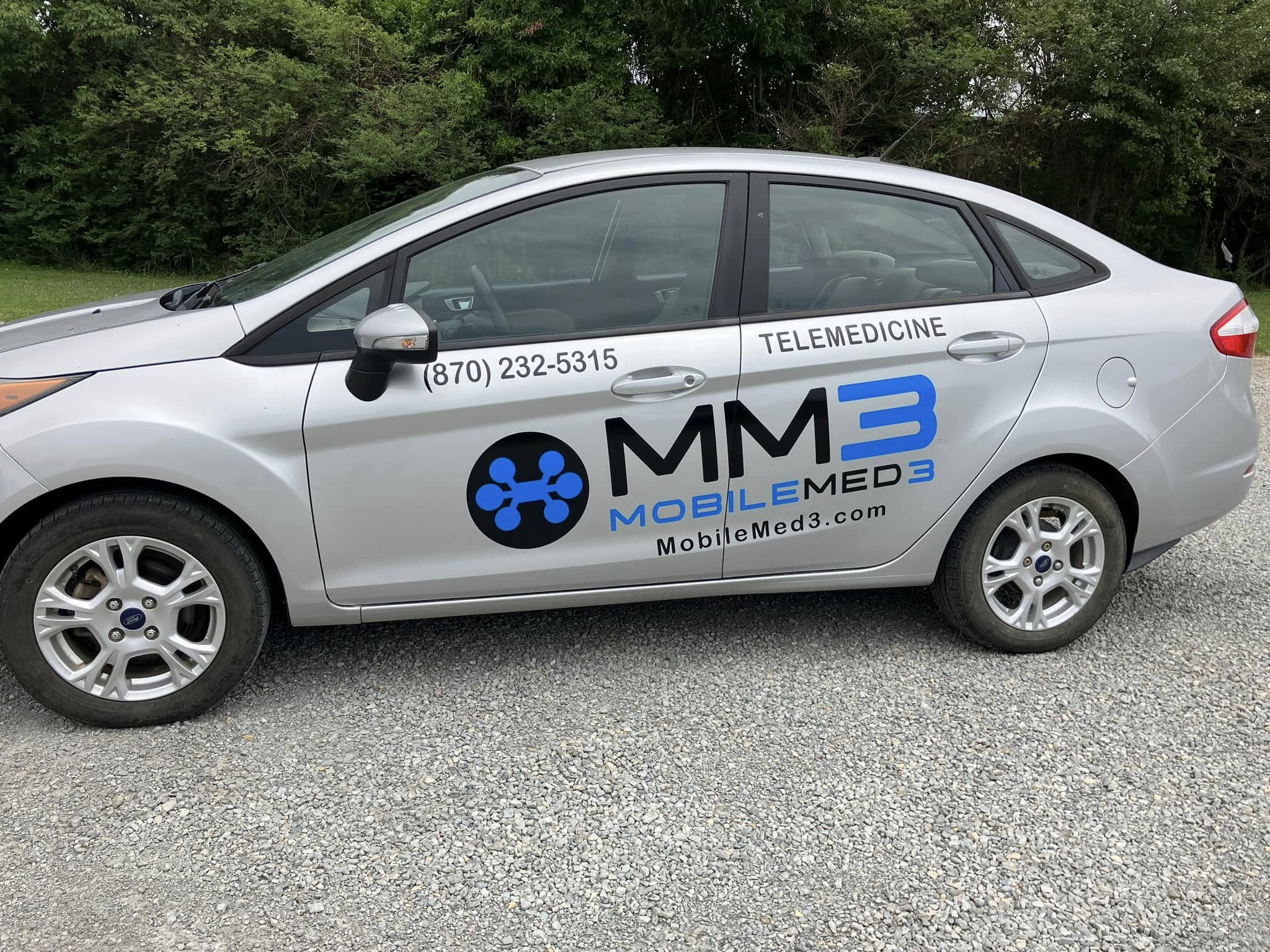 Silver Ford Fiesta with vinyl graphics for MobileMed3, a telemedicine company, parked on gravel.